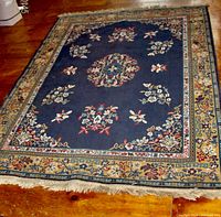 Full view of the oriental style woven rug showing its blue dominant center, floral patterns, decorative borders, and fringe ends on wooden floor.