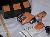 Photo showing the RIDGID cordless drill, battery charger, carrying case, and a user manual with a ruler for scale.