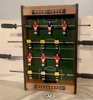 Wooden table top foosball game with plastic players in red, yellow, white and blue on metal rods with black handles, wooden frame and manual score counters.