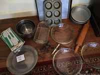 Wide view of baking lot items on floor showing glass pie plates, wooden rolling pin, metal bread pans, muffin pan, cake pans, nesting mixing bowls, metal cookie cutters, and boxed spatulas.