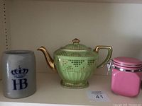 Photo showing a gray HB German beer stein, green gold trimmed teapot and pink jar