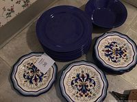 Stack of 9 solid blue dinner plates, 7 scalloped luncheon plates with floral design, and 2 small solid blue bowls.