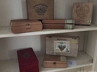Seven empty cigar boxes of various brands including Montoya, CAO Gold, Joya de Nicaragua arranged on white shelves showing wear and signs of use.
