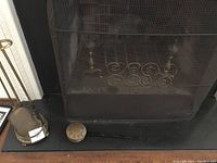 Photo showing black mesh fireplace screen with decorative scroll work and brass stand on left side holding fireplace tools including shovel and poker; vintage bed warmer resting on hearth.