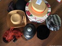 Seven hats arranged on wooden floor including straw bowler, black velvet hat, police helmet, patterned top hat and floral hat box