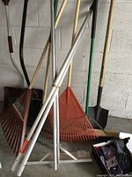 Full view of multiple lawn and garden tools leaning against a wall including rakes with plastic tines, shovels, and a white roof snow shovel.