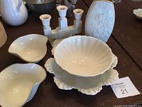 Photo showing two kidney-shaped white ceramic candy dishes, a white ceramic cocktail bowl set on a matching scalloped tray, a white candle labra with three holders, and a white ceramic vase with floral relief pattern in background.