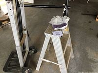 Photo showing small metal step ladder with three steps, hand dolly, and work gloves on top of ladder.