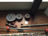Top-down view showing the assorted weight plates and two metal bars with red collars spread on the wooden floor beneath a window.