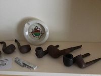 Eight pipes, metal can opener, and ceramic ashtray displayed on a shelf with a white surface and plain background.