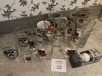 Front overview of entire lot showing assorted Princeton branded clear glasses, shot glasses, mugs, and boxed golf balls arranged on floor against floral wallpaper.