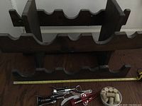 Wooden 10-slot wine rack with metal wine openers and container of used corks in front, tape measure showing about 24 inches length of rack