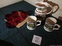 Photo showing two ceramic mugs, a ceramic pitcher with Christmas Garland design, and five red glass small drinkware glasses on a carpeted floor.