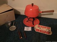Vintage orange fondue pot setup with wooden handle, lid, and drip plate, plus sterno container, drip bowl, and three fondue forks with wooden handles