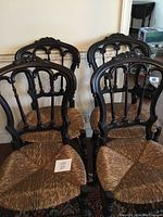 Four vintage dark wood carved shield back chairs with rush woven seats, positioned in two rows inside a room with carpeted floor.