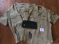 Two vintage US Army tan short-sleeve shirts with patches and insignia, green pants, green garrison cap, and four pocket tools, displayed on wooden floor.