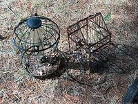 Metal round bird feeder and square bird cage placed on ground outdoors, visible pine needles around them.