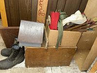 Photo showing wood storage chest, wooden bird house, cardboard box with camping gear including belts, hat, and fishing rods, and pair of black boots on floor against wood panel wall.