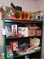 Black shelving unit with Thomas Kincade lighthouse collectible, copper-colored decorative plates, boxed cat planter, assorted cookbooks and magazines, candle holders and small decor items