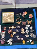 Full view of all costume jewelry items laid out on dark cloth, showing variety of pins, earrings, rings, and faux pearl necklace in box.