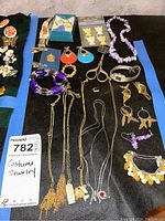 Spider web style costume jewelry laid out on black fabric with blue tape edges including earrings, necklaces, bracelets, and a watch; various colors and materials visible.
