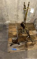 Photo showing the fireplace tool stand with broom and poker alongside two owl andirons on a wooden pallet in a concrete-floored room.