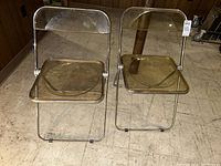 Two lucite folding chairs side by side on a tiled floor, showing transparent seat and backrest with chrome frames.