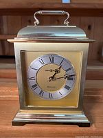 Front view of brass-tone Howard Miller mantle clock with gold and silver-tone face showing Roman numerals and ornate black hands.