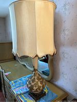 Full view of vintage table lamp with scalloped cream fabric lampshade, visible floral pattern on lamp body, ornate metal base on dresser.