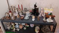 Photo showing a collection of vintage salt and pepper shakers, figurines, ceramics, glassware, and souvenirs arranged on a metal shelving unit against floral wallpaper.