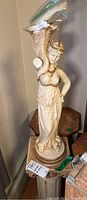 Full view of the decorative statue mounted on a wooden pedestal. The statue is a woman holding a vessel over her head, taped at the broken arm for protection.