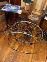 Round metal plant stand with six circular holders and other plant stands around it on wooden floor.