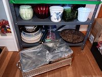Overall view of shelf with various plant pots, souvenir plates, a tapestry, and brown marbled plate on bottom shelf.