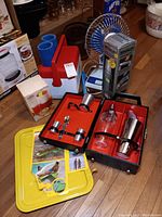 Photo showing entire lot including Stanley thermos in box, blue plastic cups in red and white cooler, small blue electric fan, boxed ice grinder, travel cocktail bar set in open black case with red interior, and yellow folding trays with vintage inserts.