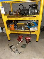 Tools and vices on yellow plastic shelving unit showing various items including two red vices, hand drill, and grinder.