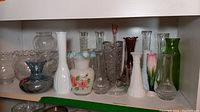 Photo of a shelf displaying twelve glass and crystal vases of various styles including ruffled edges, hand-painted floral designs, clear crystal cut patterns, milk glass, red glass and green glass vases.