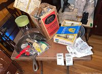 Lot displayed on wooden table including boxed Sunbeam meat grinder with wooden handle, vintage cast iron pan, plastic and metal kitchen tools, and decorative cloth napkins