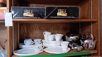 Shelf display showing vintage luncheon cup and plate sets in floral and embossed grape designs, metal teapot, creamer, sugar bowl, Penguin ice bucket, and boxed wine glass sets on top shelf.