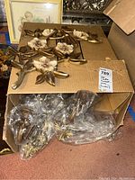 Multiple gold-tone floral plaques with cream-colored flower details inside a cardboard box, with some wrapped in plastic.