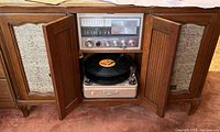 Wood cabinet with two center doors open revealing built-in metal-tone turntable with vinyl record, radio tuner control panel, control knobs.