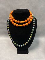 Front view of two necklaces displayed on black bust stand. The inner 24 inch necklace has round cloisonné enamel beads with floral patterns and an orange cloisonné clasp. The outer 28 inch Tibetan style necklace features carved wooden beads in various shapes including round and elongated cylinders.