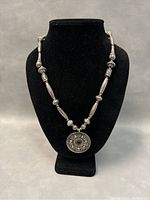 Silver-colored metal beaded necklace with large round ornate pendant on black stand