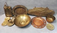Nine assorted brass items arranged on a gray surface including trays, bowls, a candle holder, and plaques.