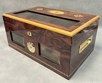 Front and side angled view of dark wood grain humidor box with beveled glass windows on front and top, round hygrometer in center front, marquetry decorative inlay on top surface, and brass side handle.