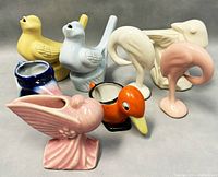 Vintage ceramic bird figurines arranged on a gray background including pink bird flower pot, blue bird creamer, white swan, orange and black bird creamer, and pink flamingo.