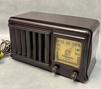 Photo of vintage brown Bakelite tube radio, showing front angle with speaker grille and tuning dial with two control knobs, power cord visible.