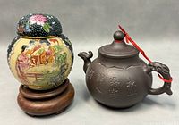 Two vintage Chinese items side by side: Yixing teapot with serpent handle featuring engraved characters and a red string on the lid, and a colorful Satsuma lidded ginger jar with floral and figurative painted decoration on a wooden base.
