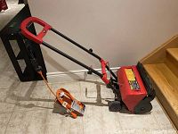 Red and black electric snow shovel with black and red handle, standing on tile floor next to stairs, orange extension cord coiled on the floor.