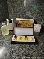 Photo showing gold tone evening bag in box, CK cologne bottle, boxed CK cologne, Dior Miss Dior boxed perfume, and a three-bottle mini cologne set in a display case.