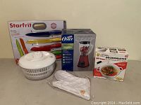Front view of all items showing Starfrit knives, Oster blender box, salad spinner, rice cooker box, and gloves